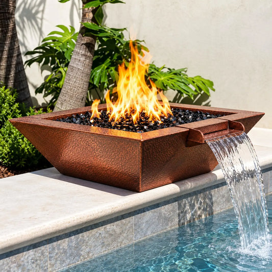 Flame Havens Cabana Hammered Copper Fire & Water Bowl displayed on a light stone ledge beside a tropical swimming pool. Tall yellow-orange flames rise through glossy black fire glass as a wide copper scupper creates a smooth waterfall into the blue water. The bowl's distinctive square design, flared rim, and rich hammered copper texture stand out against lush tropical foliage, palm trees, and natural stone pool finishes.