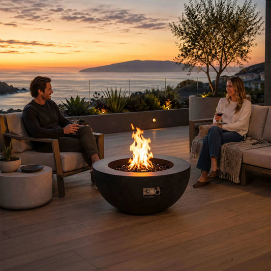 Lifestyle image of the Flame Havens Refuge Series 36-inch Round Natural Gas GFRC Concrete Fire Pit in Black burning with premium tempered black reflective fire glass on a luxury outdoor patio overlooking the ocean at sunset. Two people relax with drinks around the fire, demonstrating how the modern GFRC concrete fire pit creates a warm and inviting gathering space for outdoor entertaining.