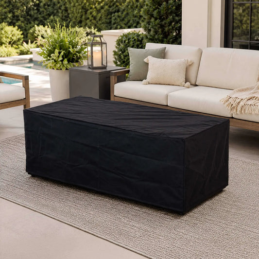 Black canvas fabric cover fitted over the Flame Havens Refuge Series 68 x 30 x 24-inch rectangular GFRC concrete propane fire pit on a refined outdoor patio with cushioned seating, decorative pillows, greenery, a lantern, and a swimming pool in the background.