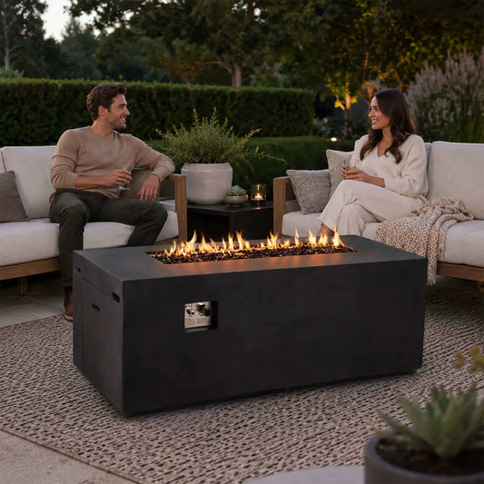 Couple relaxing on outdoor sofas beside a lit Flame Havens Refuge Series 68 x 30 x 24-inch rectangular black GFRC concrete propane fire pit. Flames rise across tempered black fire glass while the front controls and hidden tank drawer blend into the modern patio setting.