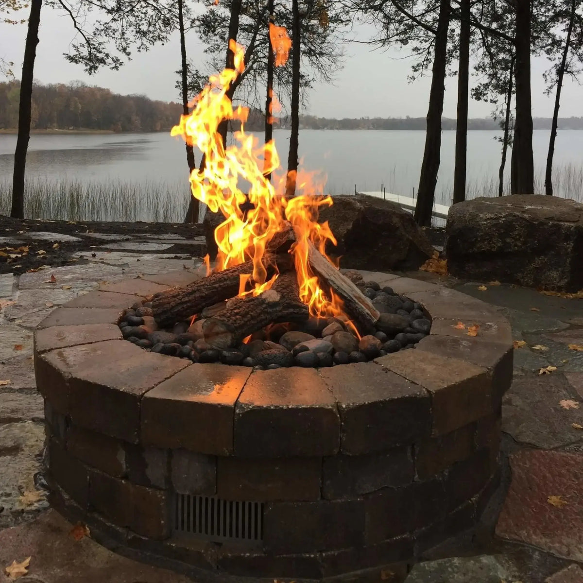 HPC Fire Hearth Products Controls Arizona Weathered Oak gas fire pit logs displayed in a real outdoor setting, burning with tall orange flames inside a circular stone fire pit near a lakeside. The logs feature realistic bark texture and charred detailing, arranged over lava rock, showcasing a natural wood-burning look ideal for backyard fire pit installations and outdoor living spaces.