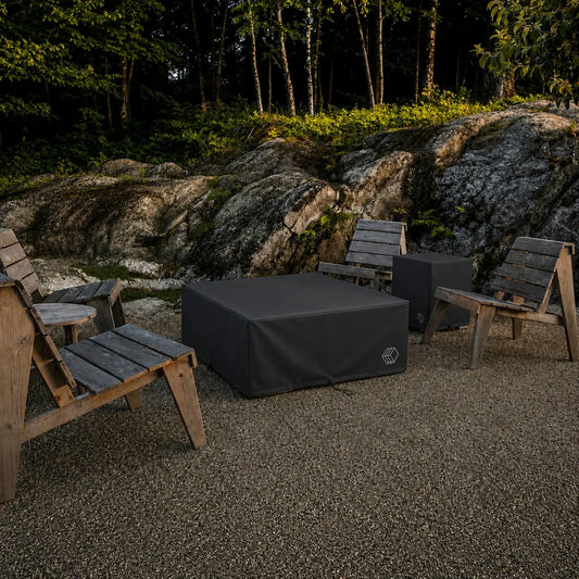 HPC Fire Hearth Products Controls Dekko GFRC Concrete Propane Tank Cover Weather Cover displayed in an outdoor gathering space surrounded by rustic wooden Adirondack chairs and natural rock landscaping. The black weather cover protects a square Dekko propane tank enclosure while blending into the backyard environment, showcasing its ability to shield outdoor fire pit accessories and propane storage components from weather, debris, and seasonal exposure.