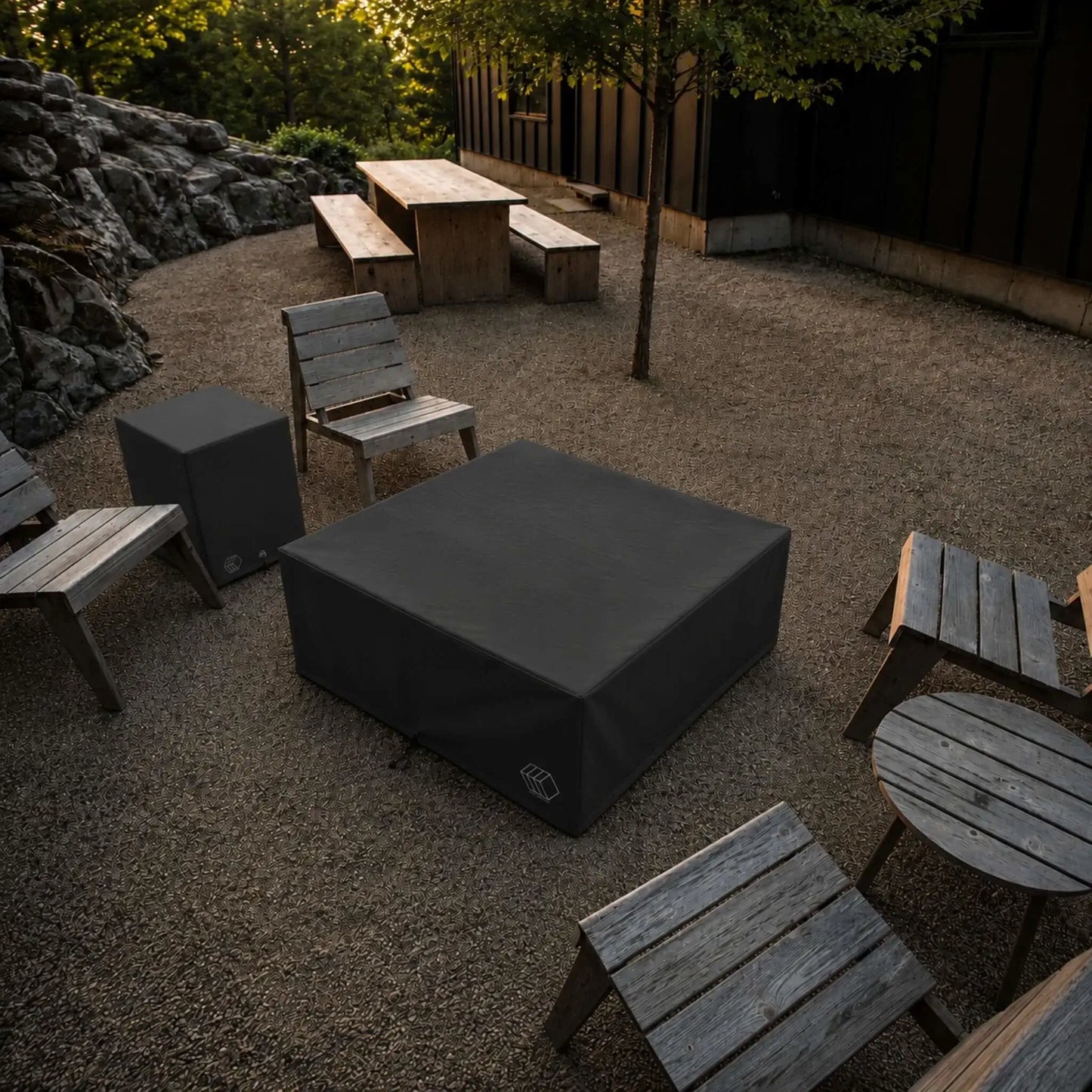 HPC Fire Hearth Products Controls Dekko GFRC Concrete Propane Tank Cover Weather Cover featured in a landscaped outdoor seating area with wooden chairs, benches, and a dining table. The fitted black protective cover encloses a square Dekko propane tank cover, demonstrating weather-resistant protection for outdoor propane storage near fire pit installations. The durable cover complements modern backyard designs while helping safeguard concrete enclosures from moisture, UV rays, and outdoor wear.