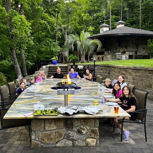 Large outdoor dining area featuring HPC Fire Hearth Products H2O n fire fire water feature integrated into a long stone table. Family and friends gather around the centerpiece where flames rise above water, combining fire and water elements for a unique luxury outdoor dining experience.