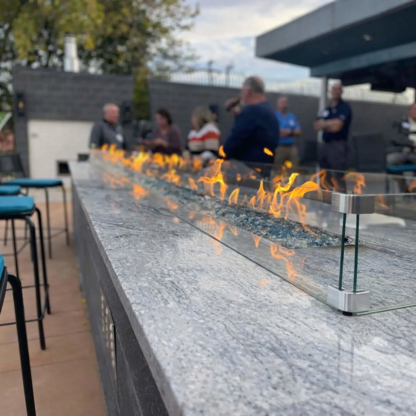 Angled close-up of a sleek linear fire pit burner filled with blue fire glass and enclosed by a clear glass wind guard. The flames reflect across the polished stone surface while a group of people stands in the softly out-of-focus background.
