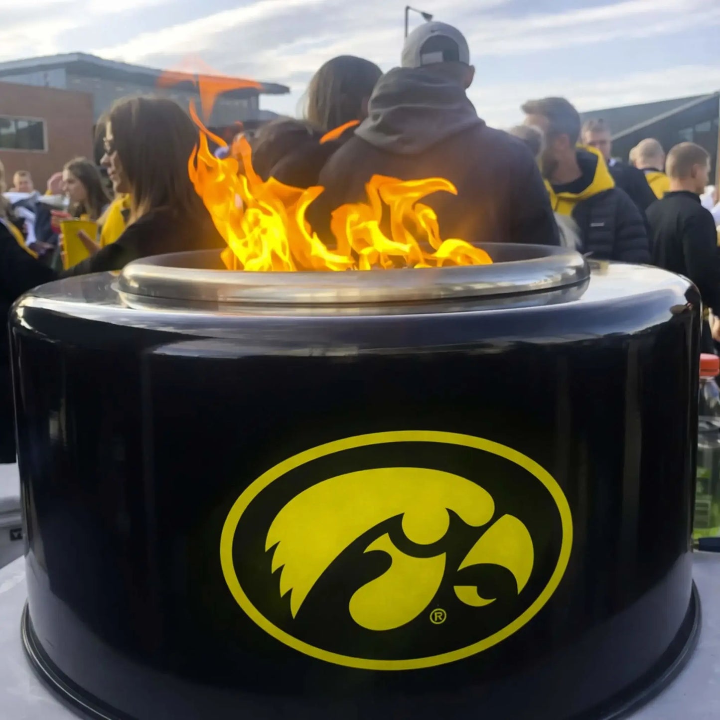 HPC Portable Sport Pit tailgate fire pit in action with Iowa Hawkeyes branding, surrounded by fans at an outdoor event. Flames rise from the top burner as people gather nearby, emphasizing the Portable Sport Pit’s role in social tailgating and outdoor entertainment settings.