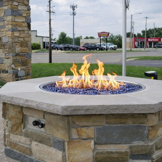 Outdoor octagon fire pit table installed in a commercial patio setting, finished with stacked stone veneer and topped with a smooth concrete cap. A vibrant gas flame burns over blue fire glass media within a round burner pan, while a push-button ignition control panel is visible on the side. The background shows a roadside environment with parked cars and nearby businesses.