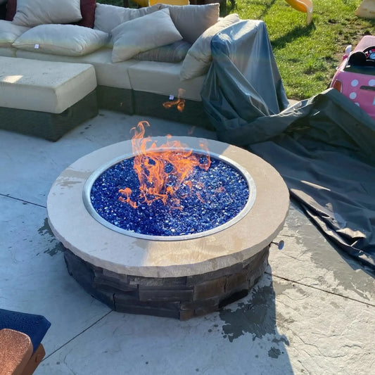 HPC Fire Inspired Hearth Products electronic ignition penta burner actively burning in a backyard fire pit filled with vibrant blue fire glass. Bright orange flames rise from the glass, surrounded by outdoor sectional seating and patio furniture, showing a cozy residential setup with strong visual contrast and modern fire feature styling.