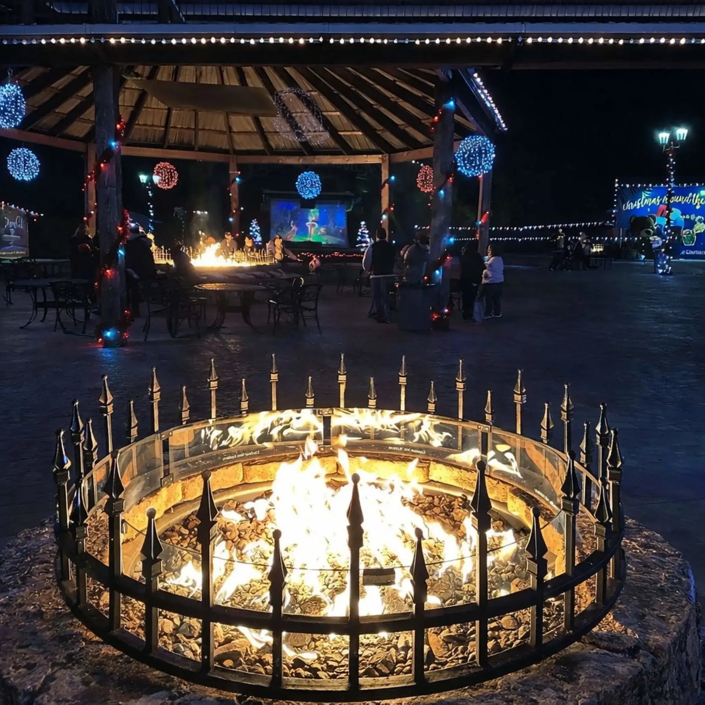 Nighttime outdoor pavilion with a circular fire pit enclosed by a decorative metal guard and glass wind barrier. The fire glows brightly as people gather nearby under string lights and festive decorations, creating a cozy, social atmosphere in a public plaza setting.