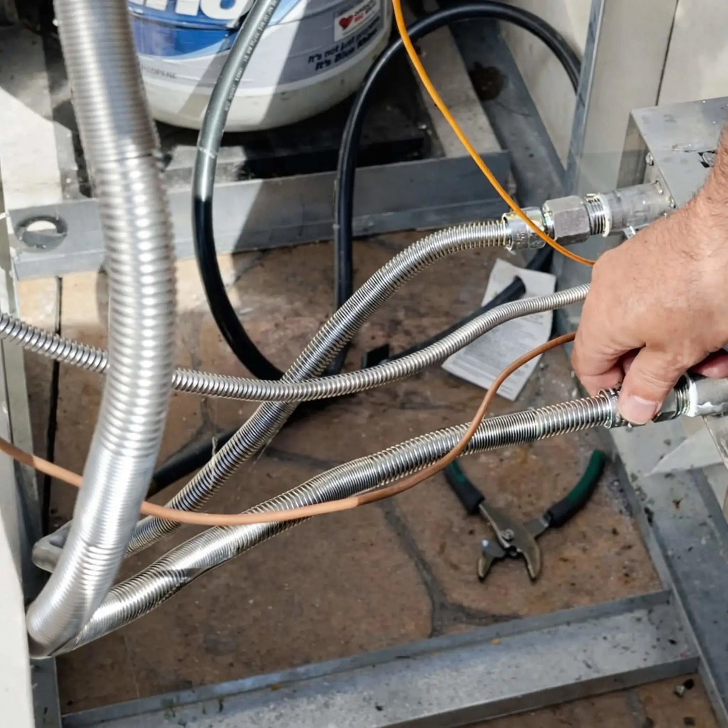 Detailed installation scene showing an HPC Fire Whistle Free stainless steel gas flex line connected to a fire pit control system and propane supply tank within a metal fire pit frame. Multiple corrugated stainless gas connectors route fuel between the valve assembly and burner components while a technician tightens the fitting by hand. Tools and wiring are visible in the enclosure, highlighting the flexible gas line’s role in high-capacity fire pit installations designed to prevent gas flow whistling.