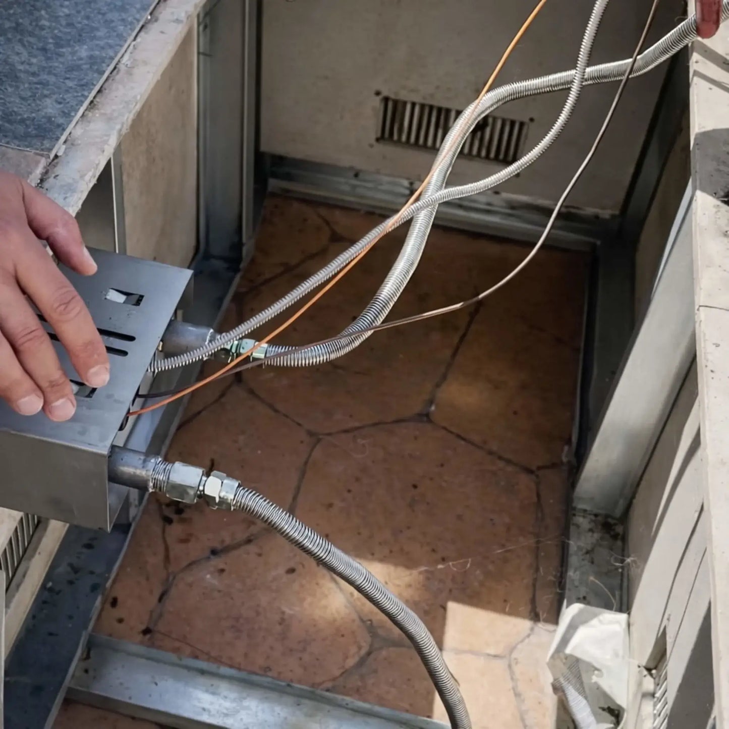Close-up lifestyle installation view of an HPC Fire Whistle Free Flex Line gas connector attached to a fire pit control system inside a metal outdoor fire pit enclosure. A stainless steel corrugated gas flex line connects the gas valve assembly to the burner system, while a technician’s hand adjusts the control box. Additional pilot and ignition wires run alongside the flex line, illustrating a professional gas fire pit installation designed to reduce gas flow noise and provide reliable fuel delivery.