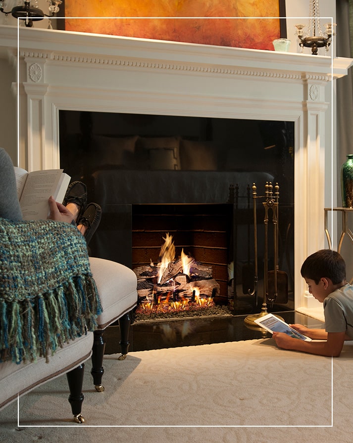 Cozy fireplace setting with a mother reading a book beside her son, who is engrossed in an article or e-book on his iPad, both basking in the comforting warmth of a gas log fireplace. The lifelike, glowing gas logs create a soothing ambiance, enhancing family bonding time in a relaxed, inviting space. This warm, intimate scene highlights the gas log fireplace as the heart of the home, offering both functionality and atmosphere for modern family living rooms.
