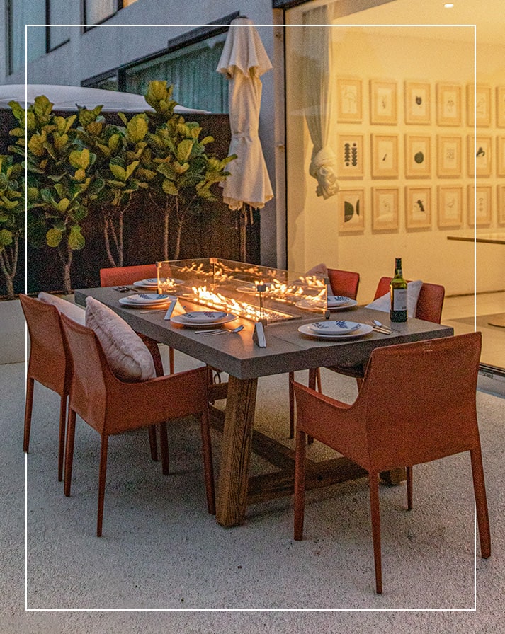 Sonoma Dining fire table by Elementi Fire set in a beautifully arranged outdoor dining area, complete with comfortable chairs, decorative pillows, wine glasses, and a dinner spread, creating an ideal setting for alfresco gatherings. The fire table combines elegant design with warmth, featuring a sleek concrete finish that complements modern and rustic decor. The lively tabletop fire adds ambiance, enhancing this inviting, stylish space perfect for memorable dining experiences under the open sky.