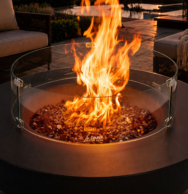 Round black fire pit with a clear glass wind guard and reflective fire glass burning on an outdoor patio.