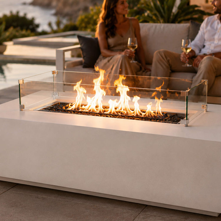 A rectangular fire pit is being enjoyed at sunset by a young couple drinking while the flame is surrounded by a glass wind guard.