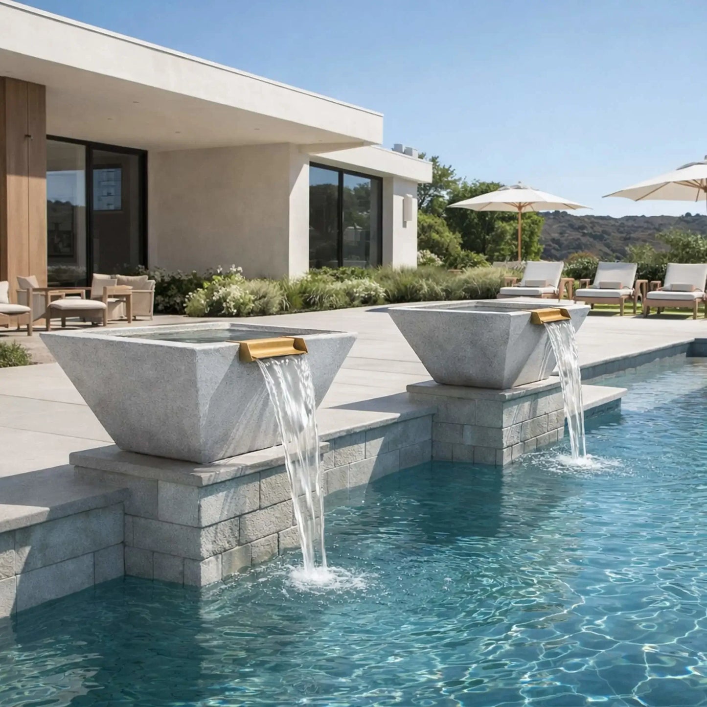 Daytime backyard pool setting showcasing gray square angular GFRC concrete water bowls placed along the pool edge on stone bases. Water flows evenly from bronze spillways into the pool, complementing a contemporary home, lounge seating, and clean modern landscaping.