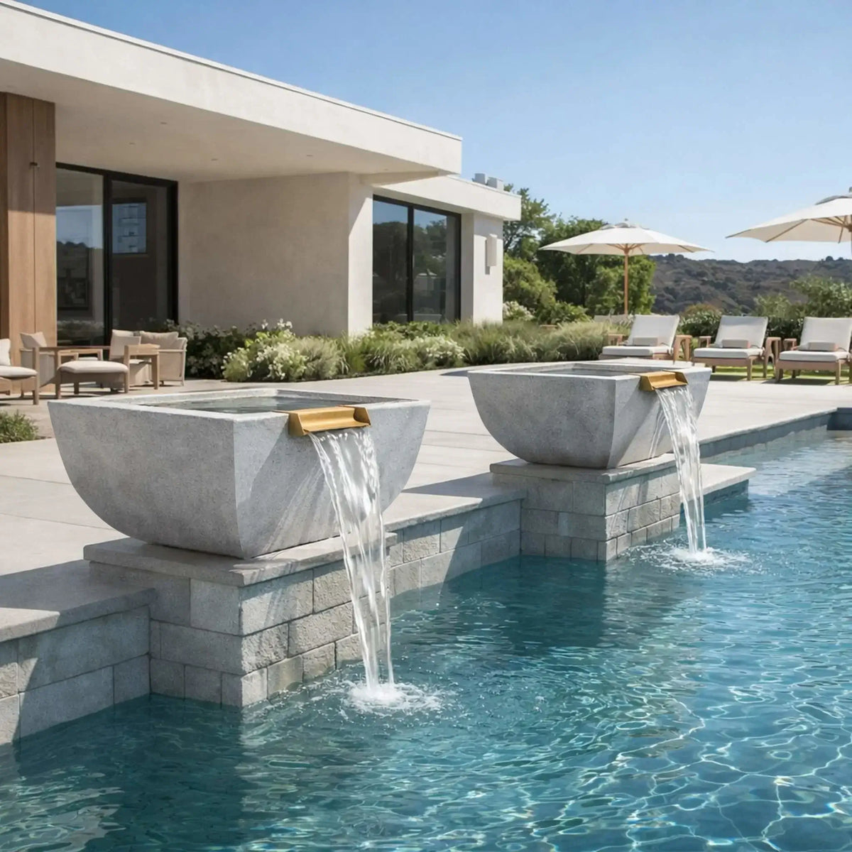 Modern poolside installation featuring two gray Serenity Series square water bowls mounted along a raised pool wall. Each architectural basin includes a copper spillway producing a smooth cascading waterfall into the pool. The contemporary backyard includes lounge chairs, umbrellas, and a minimalist home design, demonstrating a refined outdoor water feature setup.