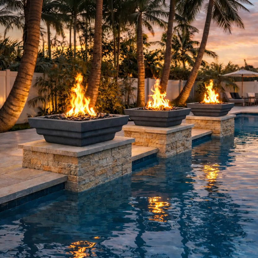 Lifestyle view of three graphite-colored Serenity Series square fire bowls installed along the edge of a modern swimming pool. Each fire bowl sits on stacked stone pedestals and burns tall natural gas flames above dark fire media. The tropical backyard setting features palm trees, warm sunset lighting, and reflections of the flames dancing across the pool water, highlighting the dramatic resort-style outdoor fire feature design.