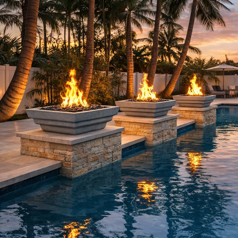 Outdoor lifestyle image showing three gray Serenity Series square fire bowls positioned on stone columns beside a luxury swimming pool. Bright flames rise from black fire glass inside each bowl, casting warm reflections on the water’s surface. Palm trees and soft sunset lighting create a tropical backyard atmosphere, showcasing the modern poolside fire feature installation.