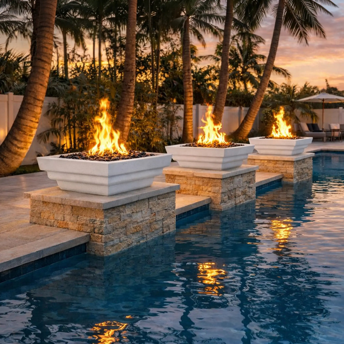 Evening poolside scene featuring three white Serenity Series square fire bowls mounted on stacked stone pedestals. Each bowl produces vibrant flames above decorative fire media, with the firelight reflecting across the blue pool water. Palm trees, warm sunset tones, and outdoor lounge furniture in the background create a luxurious tropical backyard fire feature setting.