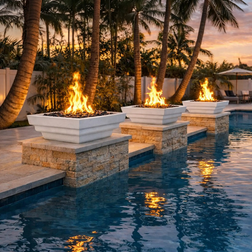 Evening poolside scene featuring three white Serenity Series square fire bowls mounted on stacked stone pedestals. Each bowl produces vibrant flames above decorative fire media, with the firelight reflecting across the blue pool water. Palm trees, warm sunset tones, and outdoor lounge furniture in the background create a luxurious tropical backyard fire feature setting.