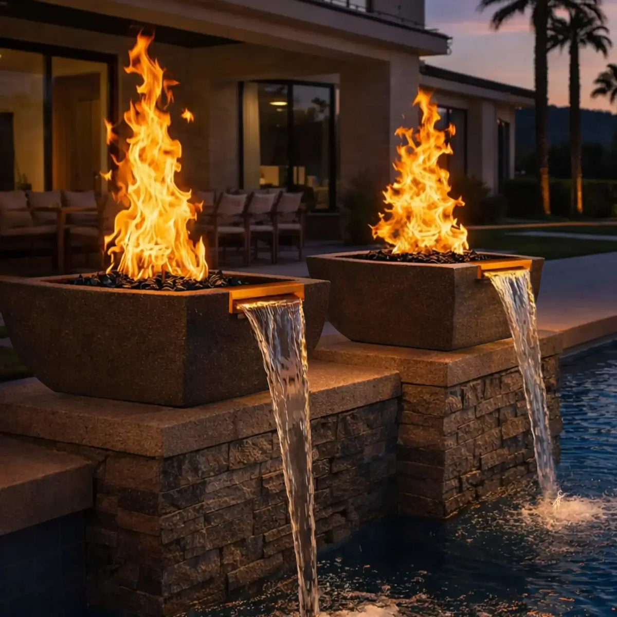 Outdoor evening patio scene featuring two black Serenity Series fire and water bowls installed on a stone pool wall. Tall flames rise above dark fire glass while copper spillways create steady waterfall streams into the pool below. A modern home and patio seating area appear softly lit in the background.
