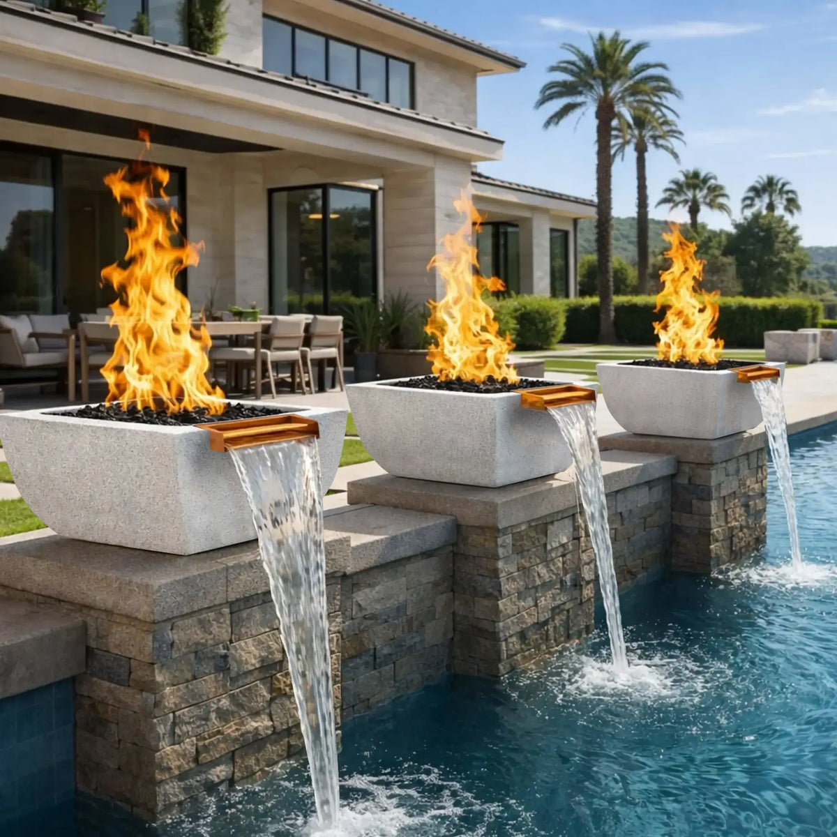 Luxury poolside scene with two gray Serenity Series fire and water bowls mounted along a raised stone wall beside a swimming pool. Tall flames burn above black lava rock while copper spillways create flowing waterfalls into the pool below. A modern home and patio seating area appear in the softly lit evening setting.