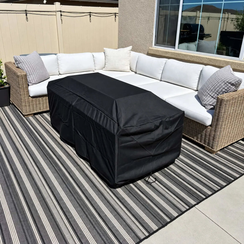 Black replacement fabric cover fitted over a Flame Havens Refuge Series 68 x 30 x 24-inch rectangular propane gas fire pit, with visible seams, fabric folds, and a drawstring at the base. The covered fire pit sits on a gray striped outdoor rug in front of an L-shaped wicker sectional with white cushions and gray throw pillows. A cream fence with string lights and a stucco wall with a large window surround the sunny patio.