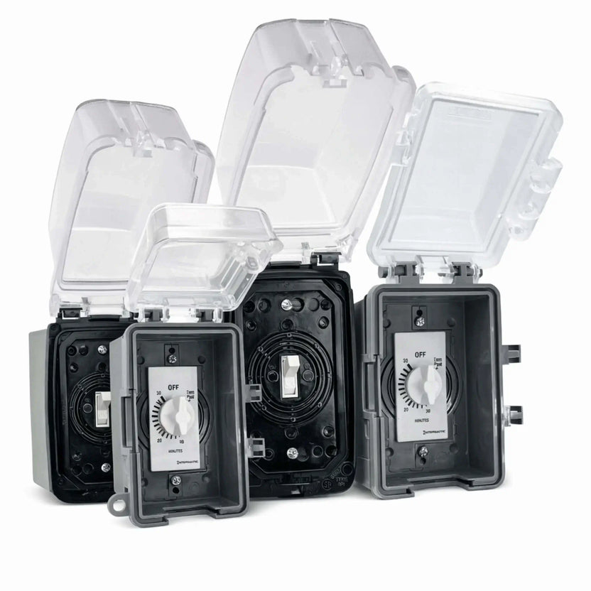 Close-up product image of American Fire Products electronic ignition control units housed in weatherproof outdoor-rated wall boxes with clear hinged protective covers flipped open, revealing integrated wall switches and 30, 60, and 120-minute countdown timer dials. The black and gray control boxes are shown side-by-side against a clean white background, highlighting the durable gasket-sealed enclosures, rotary timer knobs labeled in minutes, and heavy-duty construction designed for outdoor fire pit.