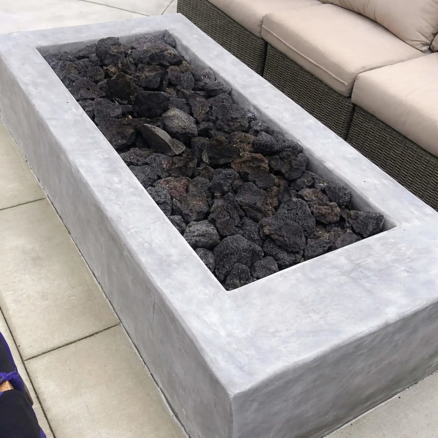 Contemporary linear concrete fire feature loaded with large lava rocks before ignition, positioned beside outdoor wicker seating on a clean patio setting.