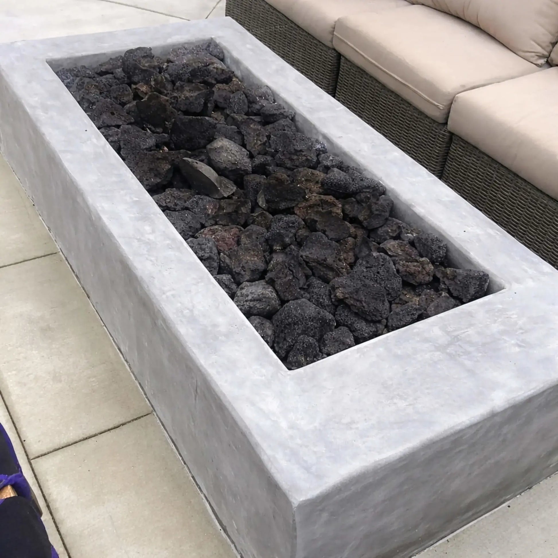 Contemporary linear concrete fire feature loaded with large lava rocks before ignition, positioned beside outdoor wicker seating on a clean patio setting.