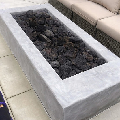 Contemporary linear concrete fire feature loaded with large lava rocks before ignition, positioned beside outdoor wicker seating on a clean patio setting.