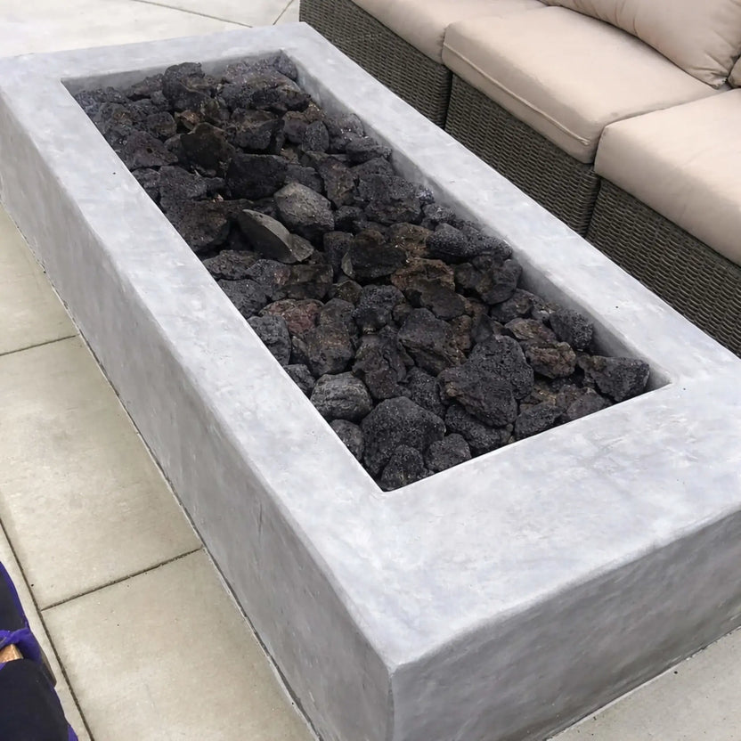 Contemporary linear concrete fire feature loaded with large lava rocks before ignition, positioned beside outdoor wicker seating on a clean patio setting.