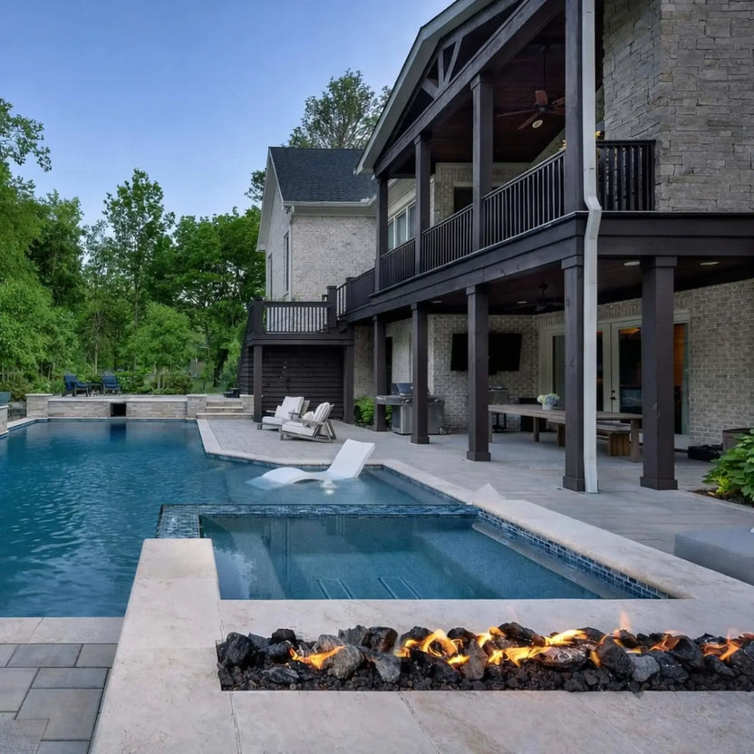 Modern luxury backyard with a custom linear concrete fire pit filled with extra extra large XXL lava rock, flames stretching across the burner in front of a resort-style pool and covered two-story patio with dark wood beams.