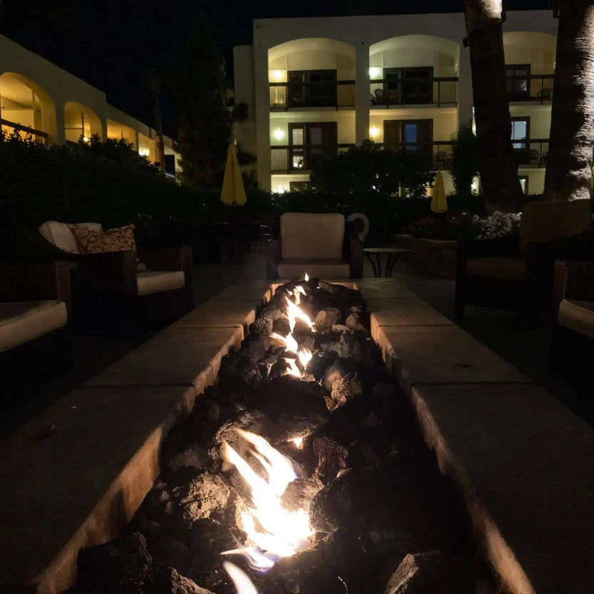 Nighttime courtyard scene featuring a long rectangular concrete fire pit packed with oversized XXL lava rock, glowing flames illuminating surrounding lounge chairs and Mediterranean-style balconies.