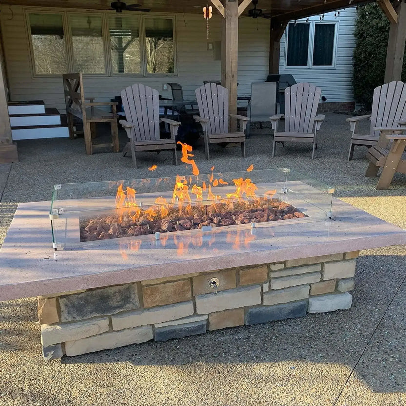 Rectangular custom stone fire table with extra extra large lava rock media and a clear tempered glass wind guard, bright flames rising evenly along the linear burner beneath a covered backyard patio.