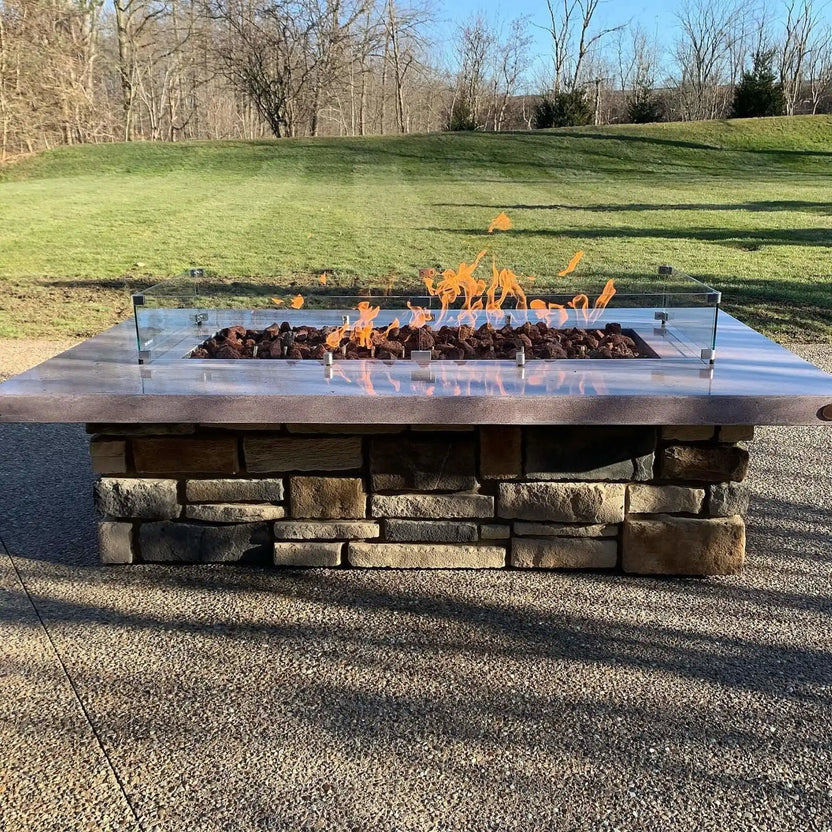 Outdoor linear concrete fire pit topped with a tempered glass wind guard and filled with chunky XXL lava rock, flames reflecting off the glass panels in an open lawn setting.