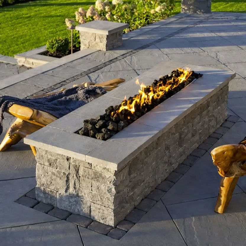 Custom rectangular concrete fire pit filled with oversized lava rocks and bright linear flames, set on a modern patio with wood seating and landscaped backyard surroundings.
