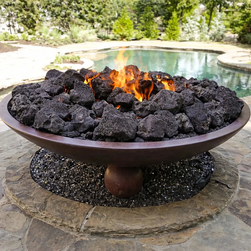 Round metal fire bowl filled with extra extra large lava rocks burning with vivid orange flames, placed on a stone patio near a backyard pool and greenery.