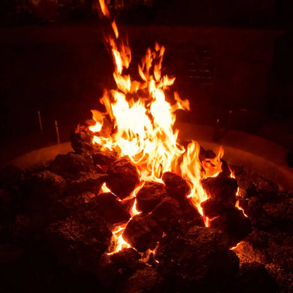Covered patio with a circular concrete fire feature surrounded by Adirondack chairs, XXL lava rock centered in the burner area with flames creating a warm focal point beneath natural wood beams.