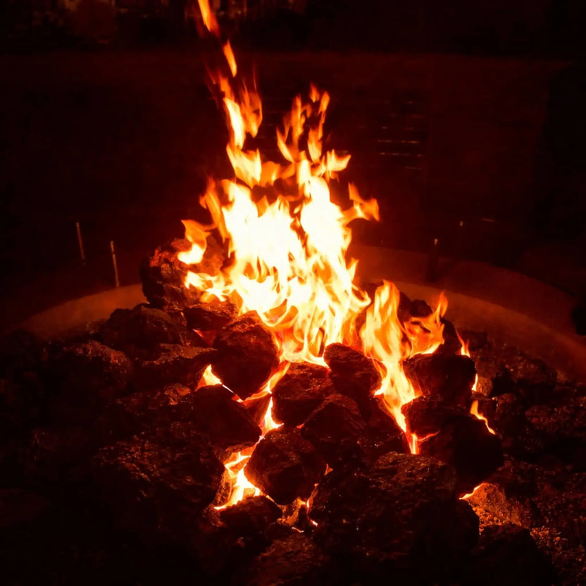 Covered patio with a circular concrete fire feature surrounded by Adirondack chairs, XXL lava rock centered in the burner area with flames creating a warm focal point beneath natural wood beams.
