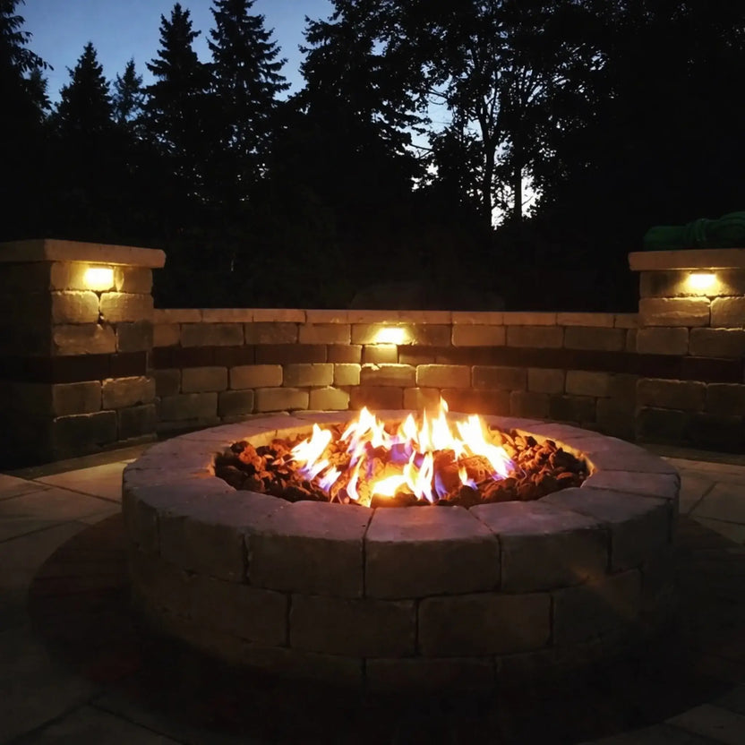 Evening shot of a round stacked-stone fire pit filled with extra extra large lava rock, blue and orange flames glowing against landscape lighting and dark tree silhouettes.