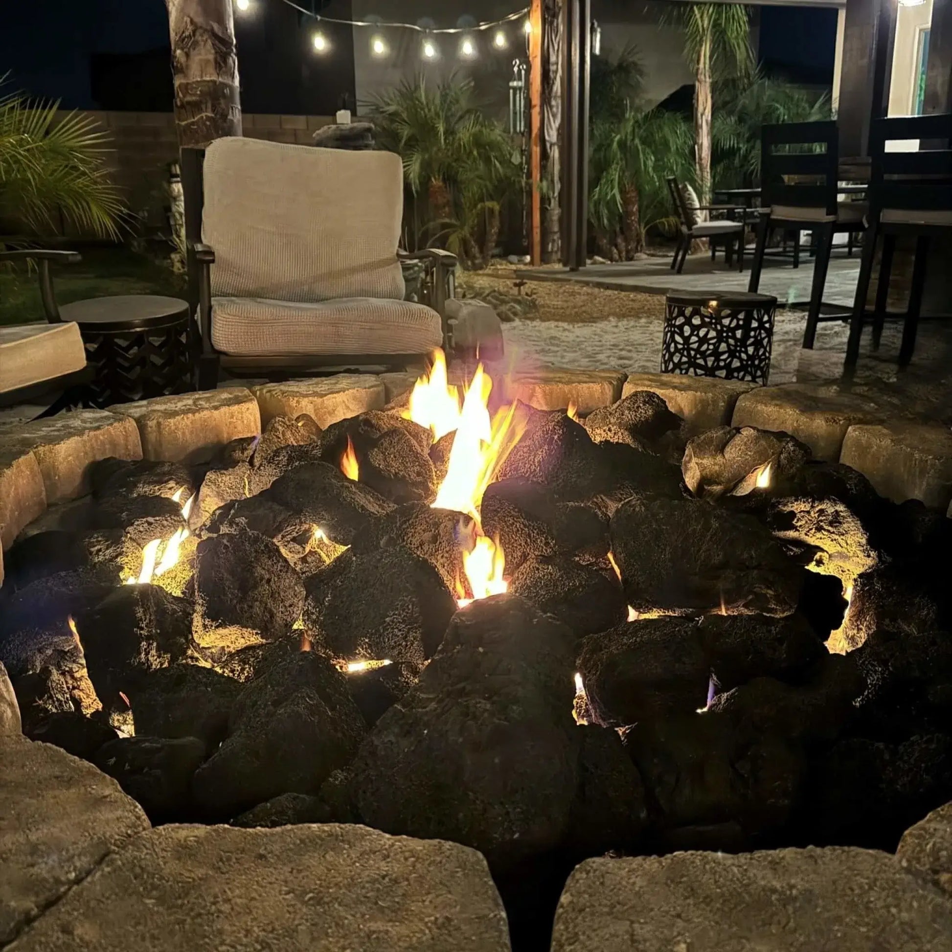 Round custom paver fire pit loaded with extra extra large lava rock, steady flames flickering at dusk against a backdrop of illuminated stone seating walls and tall evergreens.