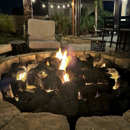 Round custom paver fire pit loaded with extra extra large lava rock, steady flames flickering at dusk against a backdrop of illuminated stone seating walls and tall evergreens.