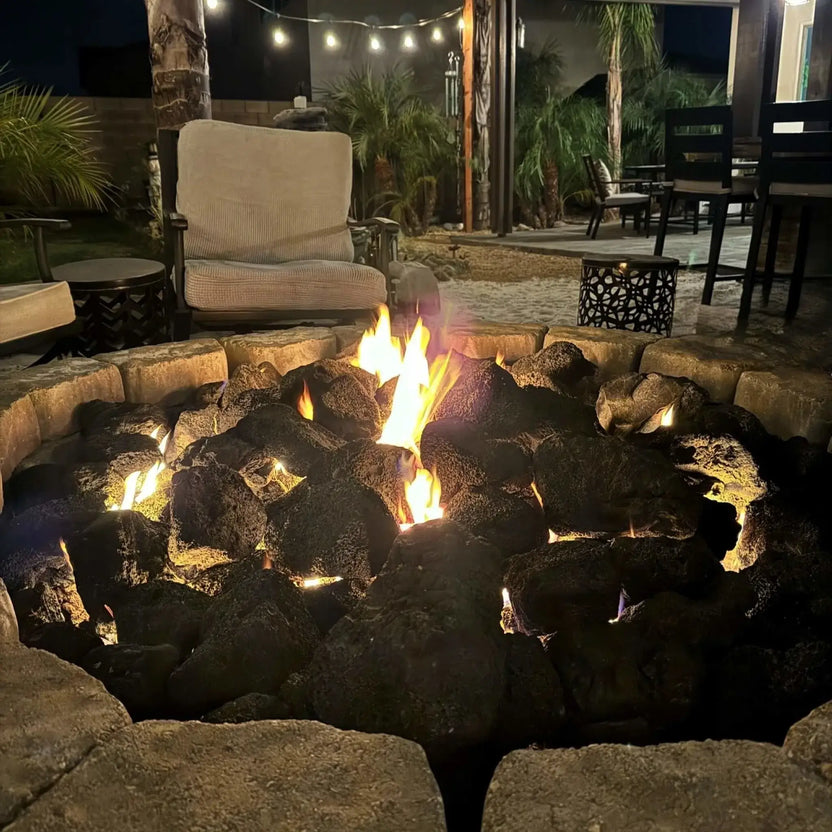 Round custom paver fire pit loaded with extra extra large lava rock, steady flames flickering at dusk against a backdrop of illuminated stone seating walls and tall evergreens.