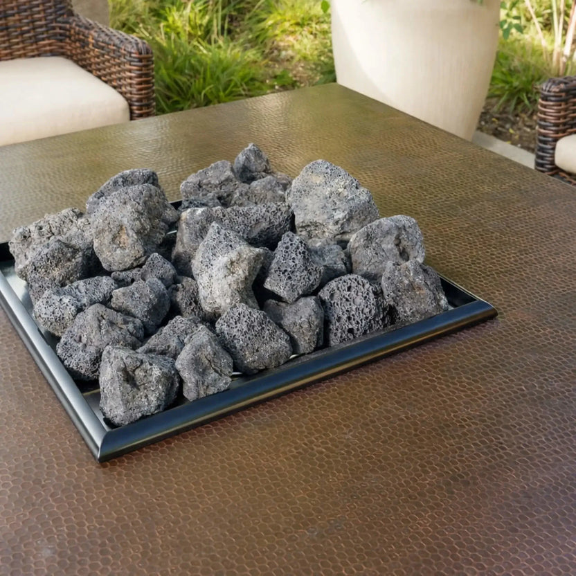 Square copper-toned fire table insert loaded with extra extra large XXL lava rock, showcasing the rugged porous texture and natural gray finish of the volcanic stones in a backyard seating area.