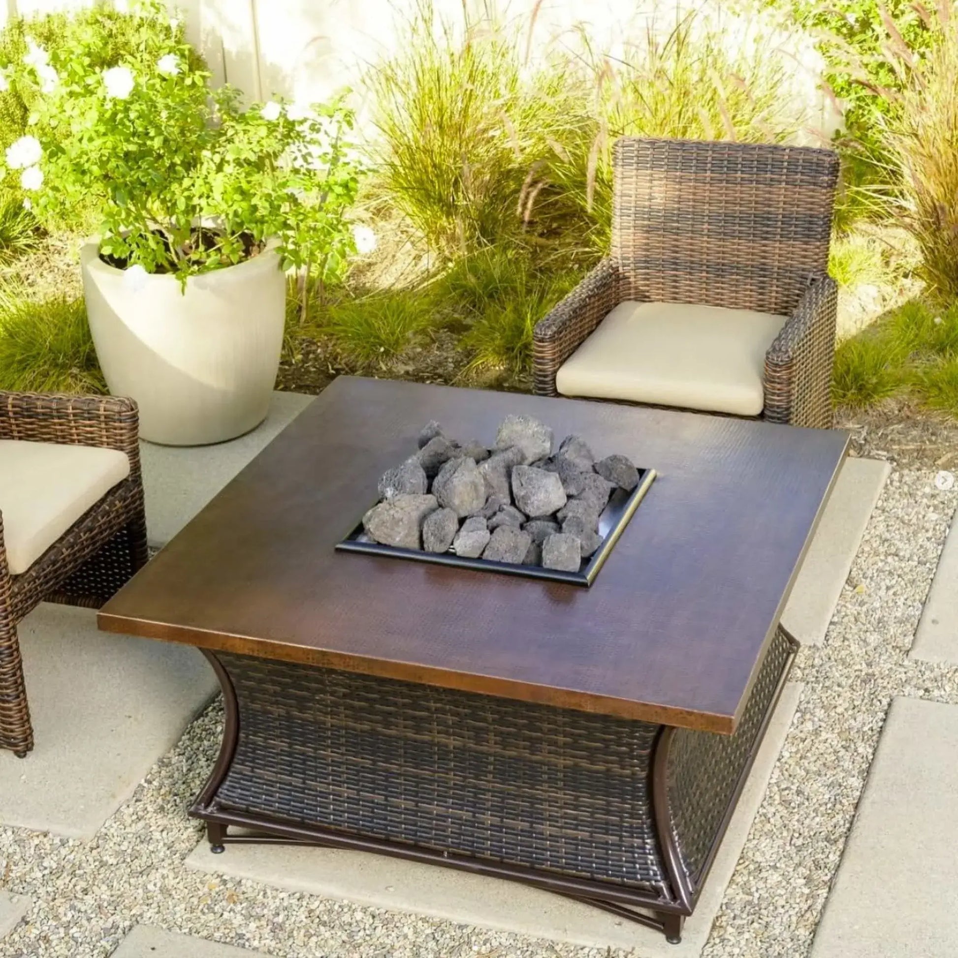 Square outdoor fire table with a copper-toned top and center tray filled with chunky lava rocks, surrounded by wicker patio chairs and decorative plants.