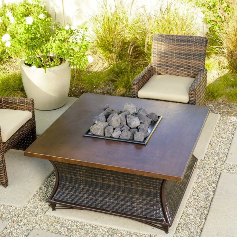 Square outdoor fire table with a copper-toned top and center tray filled with chunky lava rocks, surrounded by wicker patio chairs and decorative plants.