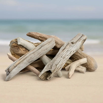 Realistic driftwood-style ceramic gas fire pit logs stacked naturally on sandy beach shoreline, featuring weathered gray and tan tones with textured grain, knots, and worn edges, set against a soft ocean backdrop for a coastal-inspired fire feature look.