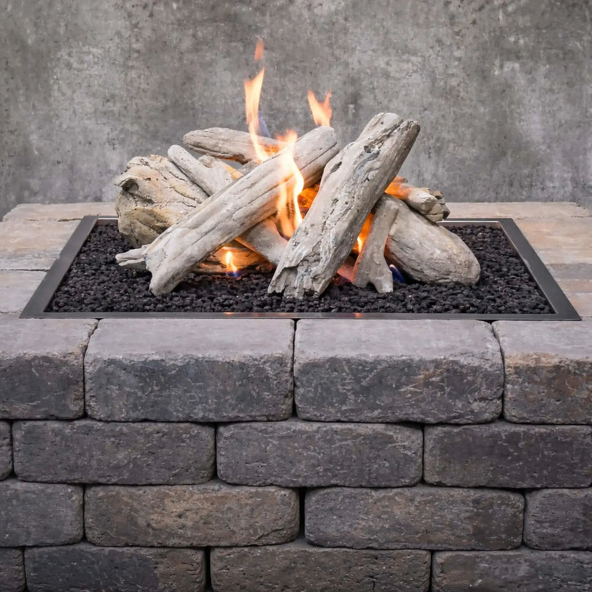 Close-up of driftwood ceramic gas fire pit logs burning over black lava rock in a rectangular burner pan, highlighting detailed wood grain texture, natural cracks, and soft gray driftwood coloring framed by a stacked stone fire pit wall.
